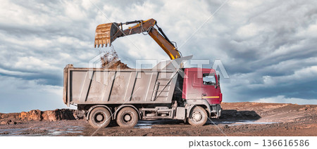 A dump truck lifts its bed to unload dirt at a construction site in a rural area. The scene shows dark clouds in the sky above the truck A dump truck lifts its bed to unload dirt at a construction site in a rural area. The scene shows dark clouds in the sky above the truck 136616586