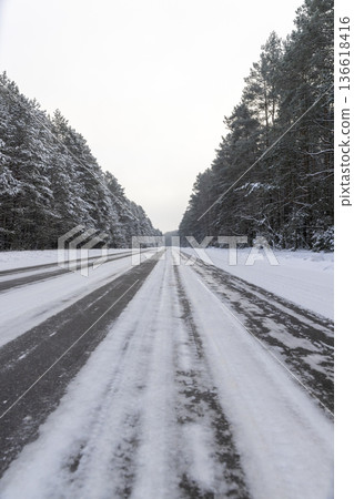 road for cars through a winter forest in the snow , a narrow track covered with snow from cars in the forest in the winter, trees grow on the side of the road in the snow in a mixed forest 136618416