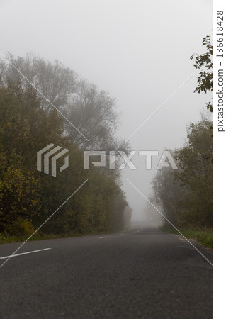 a narrow country road with yellow trees on the edge of the road in the autumn season, cloudy weather with a gray nondescript sky, thick fog in the morning due to climate change 136618428