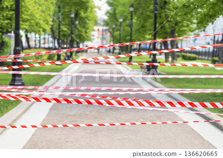 Red and white barrier tape blocking pedestrian path in city park, empty walkway, green trees in background. Concept of safety, restriction, emergency safety in city, barrier and closure, urban caution 136620665