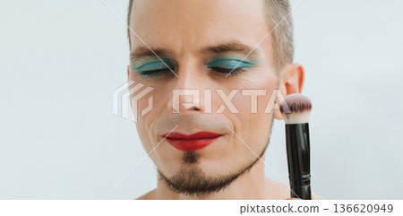 feminine gay man with makeup and cosmetics on face. Handsome adult transgender male with red lipstick on lips and bright eye shadows holds a blush brush on a white background feminine gay man with makeup and cosmetics on face. Handsome adult transgender male with red lipstick on lips and bright eye shadows holds a blush brush on a white background 136620949