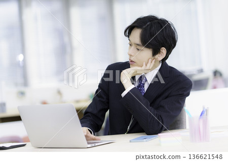 A young Japanese man in a suit with a troubled expression working on a laptop in an office A young Japanese man in a suit with a troubled expression working on a laptop in an office 136621548