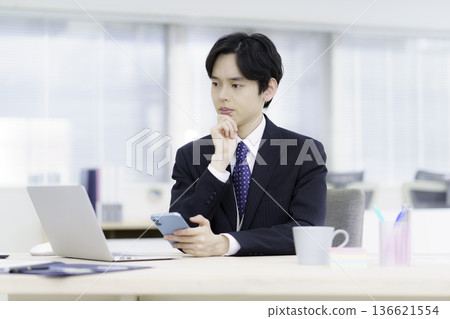 A young Japanese man in a suit with a troubled look on his face holding a smartphone in his hand in the office 136621554