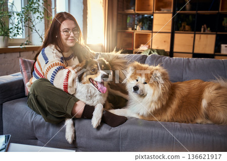 Woman sitting with two dogs on sofa in warm modern apartment. Woman sitting with two dogs on sofa in warm modern apartment. 136621917