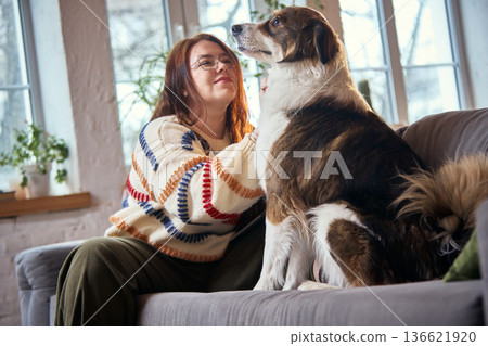 Happy dog sitting beside female owner on sofa in cozy living room. Happy dog sitting beside female owner on sofa in cozy living room. 136621920