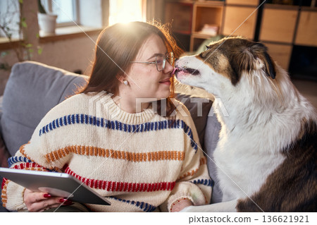 Dog licking woman face while she holds tablet on couch. 136621921