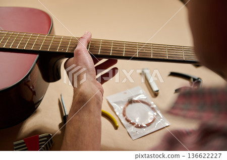 Hands adjusting and changing strings on an acoustic guitar, showing routine maintenance and repair with tools and replacement strings on a workshop table. Hands adjusting and changing strings on an acoustic guitar, showing routine maintenance and repair with tools and replacement strings on a workshop table. 136622227