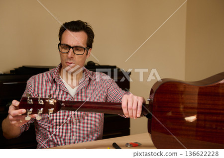 Man at a workbench adjusting and changing guitar strings, demonstrating careful instrument maintenance and repair techniques. Man at a workbench adjusting and changing guitar strings, demonstrating careful instrument maintenance and repair techniques. 136622228