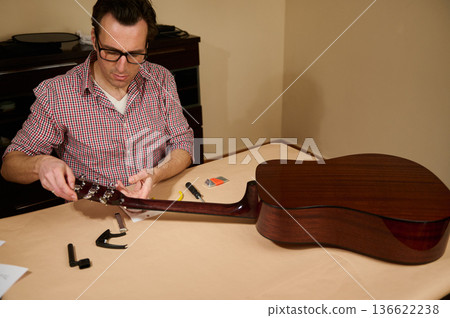 Master adjusting and changing strings in guitar, showing a man performing guitar maintenance and repair at a home workspace with tools and parts laid out for restringing and tuning. 136622238