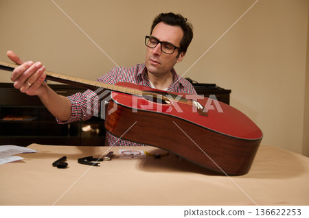 Man adjusting acoustic guitar strings on a table, demonstrating careful guitar maintenance and repair with tools and focused attention. 136622253