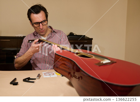 Man adjusting acoustic guitar strings at a table, demonstrating guitar maintenance, string replacement and repair techniques for instrument care. Man adjusting acoustic guitar strings at a table, demonstrating guitar maintenance, string replacement and repair techniques for instrument care. 136622255