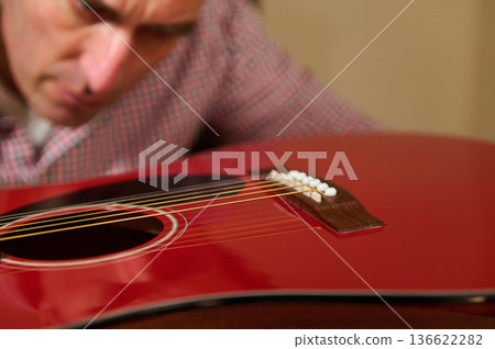 Man carefully adjusting strings on a red acoustic guitar, demonstrating hands-on guitar maintenance, string replacement and repair for musicians and instrument care. 136622282