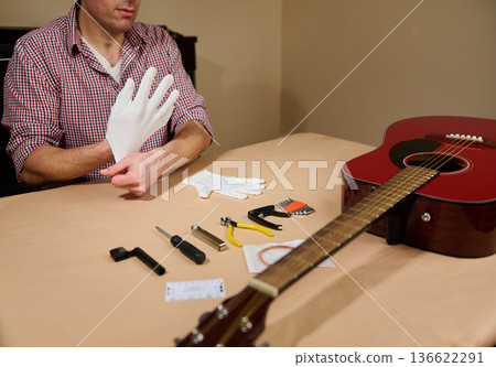 Man putting on gloves to change guitar strings with tools on a table, showing careful instrument maintenance, repair and restringing for acoustic guitar care. Man putting on gloves to change guitar strings with tools on a table, showing careful instrument maintenance, repair and restringing for acoustic guitar care. 136622291