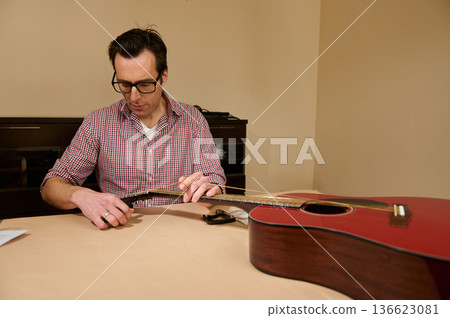 Man Changing Strings on Red Acoustic Guitar at Table - Guitar Maintenance and Repair Man Changing Strings on Red Acoustic Guitar at Table - Guitar Maintenance and Repair 136623081