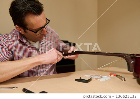 Man Replacing Guitar Strings at Table - Guitar Maintenance and Repair by Focused Musician Man Replacing Guitar Strings at Table - Guitar Maintenance and Repair by Focused Musician 136623132