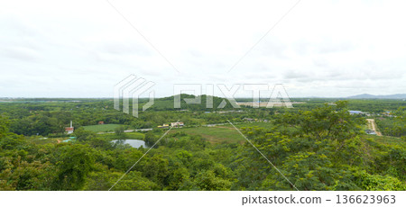 Elevated wide angle view of a lush tropical valley in Thailand with a small lake, green forests, and a white temple surrounded by trees under a cloudy sky. Panoramic scenic landscape photography. Elevated wide angle view of a lush tropical valley in Thailand with a small lake, green forests, and a white temple surrounded by trees under a cloudy sky. Panoramic scenic landscape photography. 136623963
