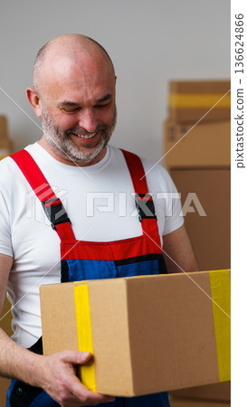 Man holds a cardboard box in a storage area while looking happy and engaged Man holds a cardboard box in a storage area while looking happy and engaged 136624866