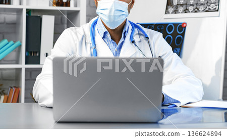 Doctor working on laptop in office while wearing mask during day Doctor working on laptop in office while wearing mask during day 136624894