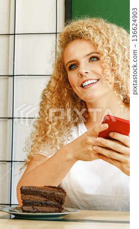 Woman smiles while holding phone near a plate of cake in a cafe 136624903