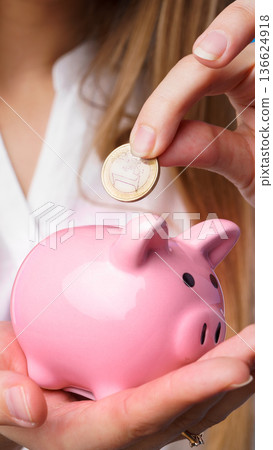 Person drops a coin into a pink piggy bank during savings activity at home Person drops a coin into a pink piggy bank during savings activity at home 136624918