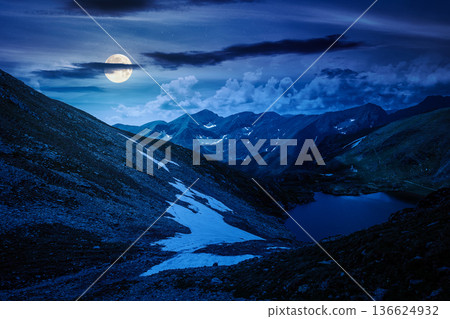 mountain lake in alpine summer landscape at night. beautiful view surrounded with clouds on a dark sky in full moon light. fresh water, snow and rocky slopes. mysterious remote fantasy highlands 136624932