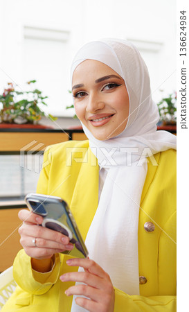 Woman in a yellow jacket using her smartphone in a bright indoor setting with plants 136624984