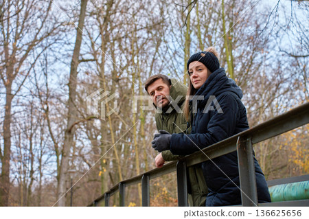 Man and woman in warm jackets standing by metal railing in park Man and woman in warm jackets standing by metal railing in park 136625656