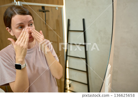 Woman standing in bathroom and applying cream on nose while looking at mirror 136625658