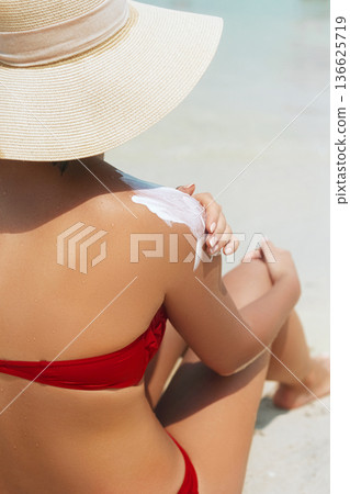 Beautiful woman in hat  sitting on the beach and applying  sunscreen spray on her body.Skincare. Body Sun protection suncream. 136625719