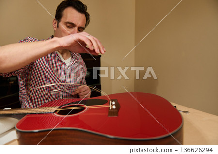 Man Changing Strings on Red Acoustic Guitar in Home Workshop Man Changing Strings on Red Acoustic Guitar in Home Workshop 136626294