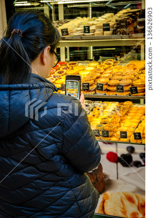 Anonymous woman in black puff jacket taking photo of traditional Portuguese pastries including famous pastel de nata in bakery window with smartphone. Food photography for social media blog. Tourist 136626903