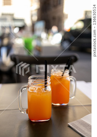 Two mason jars filled with fresh orange juice served on a table at an outdoor cafe in the historic center of Rome with blurred city street background and warm afternoon light. 136630514