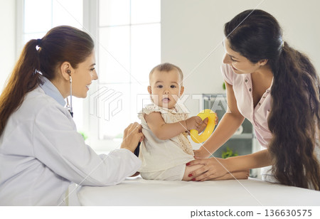 Pediatrician examining infant with mother in clinic, baby holding toy during visit 136630575