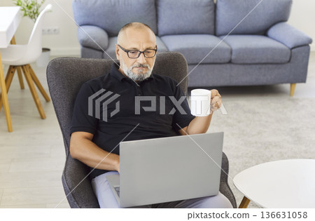 Senior man with laptop drinking coffee while working remotely in cozy home 136631058