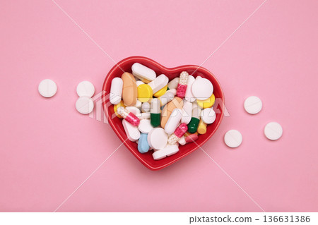 Heart-shaped bowl with a colorful assortment of various supplements on a pink background. Health, wellness, and care pharmacology concept. Heart-shaped bowl with a colorful assortment of various supplements on a pink background. Health, wellness, and care pharmacology concept. 136631386