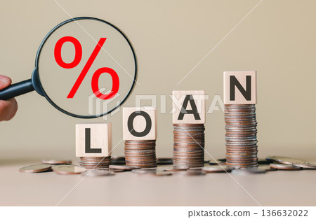 Stacked coins with wooden blocks spelling "LOAN" and a magnifying glass showing a red percentage symbol, representing interest rates, personal finance, debt and loan application process. lending money 136632022