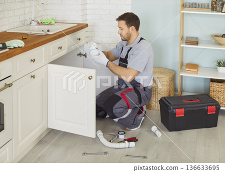 Man plumber in uniform with toolbox replacing the water filter or fixing broken pipe in kitchen. 136633695