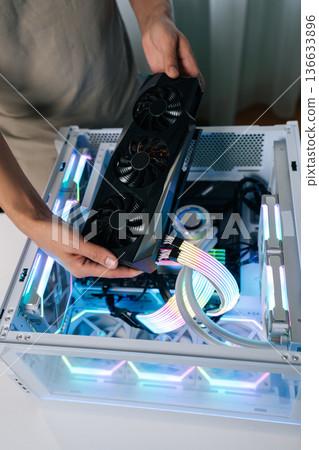 Vertical shot of man inserting new graphics processing unit into desktop computer setup with white case, visible fans, vibrant led lighting, representing hardware maintenance and technology upgrade. 136633896