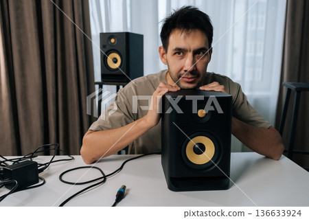 Portrait of happy young man thinking and looking away, resting arms on professional monitor speaker on white table in home studio, reflecting on creative ideas and sound. Portrait of happy young man thinking and looking away, resting arms on professional monitor speaker on white table in home studio, reflecting on creative ideas and sound. 136633924