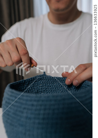 Vertical cropped shot of man enjoying creative hobby, crocheting blue yarn basket at home, hands crafting handmade item with needle and thread, engaging in relaxing and skillful activity, close-up. Vertical cropped shot of man enjoying creative hobby, crocheting blue yarn basket at home, hands crafting handmade item with needle and thread, engaging in relaxing and skillful activity, close-up. 136633925