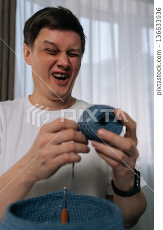 Vertical portrait of funny young man laughing and squinting with joyous emotion while engaged in relaxing hobby of crocheting, holding ball of blue yarn working on textile project with crochet hook. 136633936