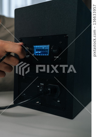 Male hand inserting balanced input cable into professional studio monitor speaker, displaying audio equalization settings on digital screen, managing sound output, close-up cropped shot. Male hand inserting balanced input cable into professional studio monitor speaker, displaying audio equalization settings on digital screen, managing sound output, close-up cropped shot. 136633957