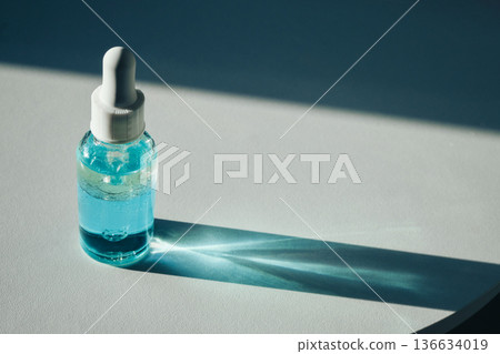 Skincare bottle with blue liquid standing on white surface casting shadow 136634019