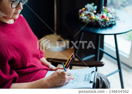 Woman drawing work-life balance wheel sitting on armchair at cozy dark home interior. Self-reflection, life planning for next new year. Personal Year review. Coaching tools. Finding Balance in Life Woman drawing work-life balance wheel sitting on armchair at cozy dark home interior. Self-reflection, life planning for next new year. Personal Year review. Coaching tools. Finding Balance in Life 136634502