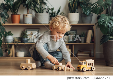 Blonde child boy sits on floor of cozy living room and playing with wooden toys, pushing small truck and stacking blocks. Indoor plants on shelves, warm atmosphere. Cute child, life, family 136634737