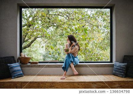 Beautiful woman sitting in front of large window, drinking tea. 136634798