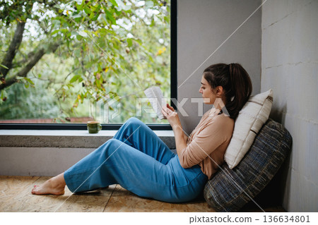 Hygge moment for beautiful woman sitting by a large window with a book. Woman is enjoying time for herself, reading. 136634801