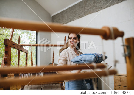 Young businesswoman next to wooden loom, holding pillows with woven covers made from recycled fabrics. Upcycling in business and circular economy. 136634824