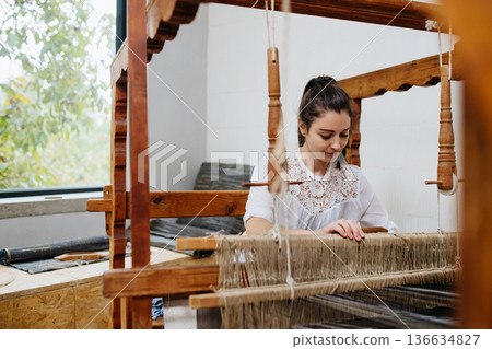 Young businesswoman sitting at a wooden loom, weaving a rug from old fabrics. Upcycling in business and circular economy. 136634827