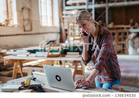 Female woodworker making phone call with client, standing in carpentry workshop. 136634830